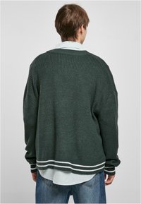 Maglione in lana di un verde scuro con una vestibilità ampia, texture a coste e dettagli a righe bianche all'orlo e ai polsini. Indossato sopra una camicia azzurro chiaro.
