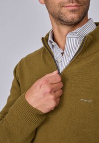 Pull en tricot côtelé vert olive avec un col zippé, porté sur une chemise blanche et grise rayée, présentant un logo brodé discret sur la poitrine.
