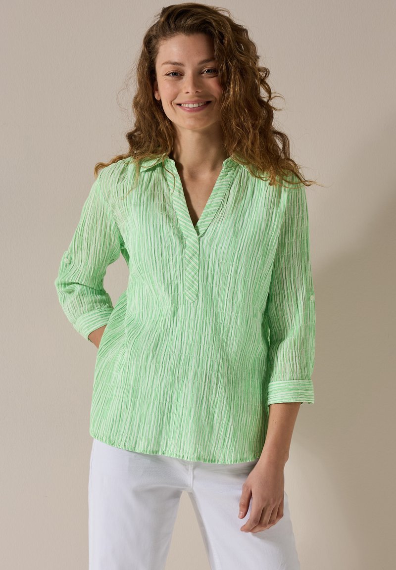 Blusa verde chiaro, con texture e righe verticali, colletto e maniche a tre quarti, abbinata a pantaloni bianchi.