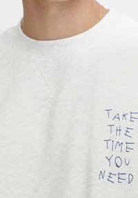 Jasnoszary sweter z teksturowanej tkaniny. Ma dekolt w kształcie litery V oraz niebieski haft na przodzie.