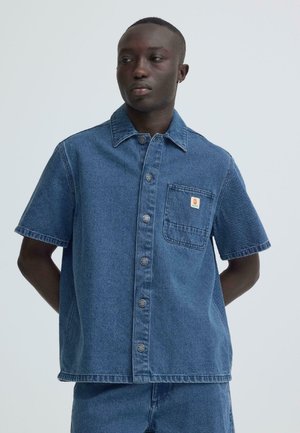 Muž ve krátkém modrém džínovém košili s knoflíky a přední kapsou, stojící se založenýma rukama za zády, dívající se stranou.