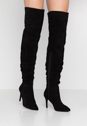 Bottes noires montantes en suede, avec un bout pointu, un haut talon aiguille, et une fermeture éclair sur le côté avec des détails froncés.