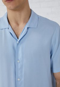 Chemise bleu clair à manches courtes avec un col camp, texture lisse et deux boutons sur le devant. Détails de couture minimalistes.