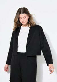 Blazer nero accorciato con chiusura a un solo bottone, rever a punte e una texture liscia, indossato sopra una camicia bianca con testo.