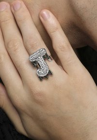 Rhinestone-besat ring formet som bogstavet "J" med grønne ædelsten, vist på en hånd med lys hudtone.
