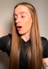 Young person with long straight hair turning head left, mouth slightly open, wearing a black shirt, visible tattoo on left arm.