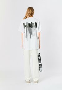 Camiseta blanca de gran tamaño con un diseño gráfico negro en la parte posterior, complementada con pantalones blancos holgados y un detalle de correa de marca.