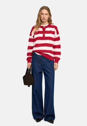 Czerwono-biały sweter w paski z okrągłym dekoltem i plisą na guziki, zestawiony z szerokimi, ciemnoniebieskimi dżinsami oraz małą brązową torebką.