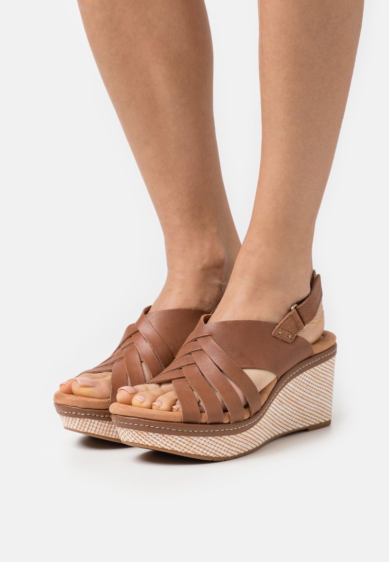 Jambes portant des sandales compensées tressées en cuir marron avec des brides à la cheville et des talons plateforme texturés sur un fond clair uni.