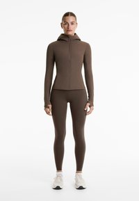 Top de exerciții lung cu mâneci, de culoare maronie, cu fermoar în față și glugă, asortat cu colanți maronii, purtați cu adidași ușori.