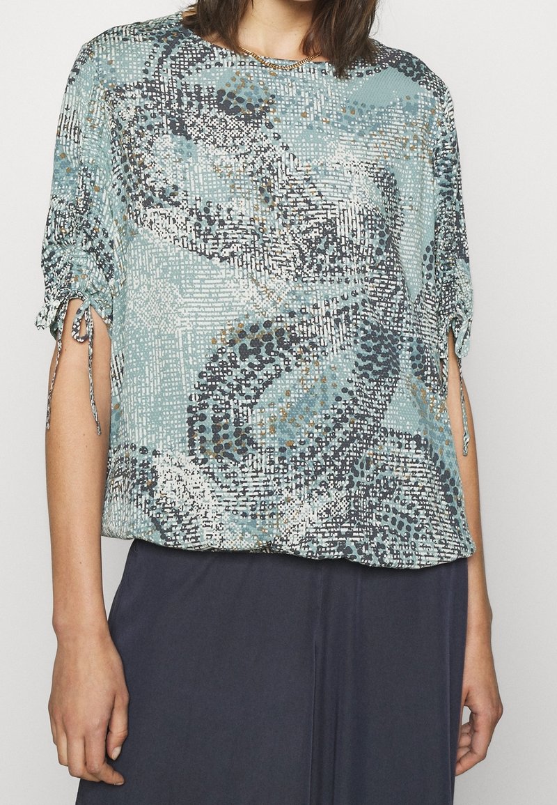 Top a maniche corte, di vestibilità ampia, in teal con motivo astratto in blu scuro, crema e dettagli dorati; presenta maniche annodate e un texture morbida.