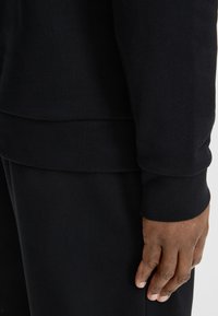 Tessuto di felpa nero a coste con una texture liscia, caratterizzato da un orlo arrotondato e polsini aderenti, mostrato parzialmente con una mano appoggiata sopra.