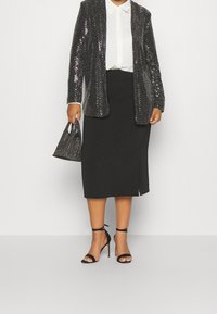 Persona che indossa una giacca nera con paillettes, una camicia bianca, una gonna nera midi e sandali neri con tacco alto, mentre tiene in mano una borsa di perline nera.