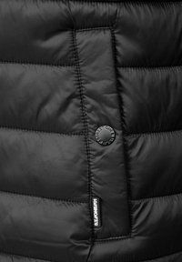 Detail černé bundy s péřovým zateplením, obsahující prošívané části, kruhový logový knoflík a značkovou etiketu na spodní části. Hladká textura látky.
