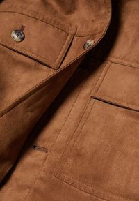 Veste en daim marron avec deux poches poitrine à rabat sécurisées par des boutons, coutures apparentes et boutonnières sur le panneau avant.