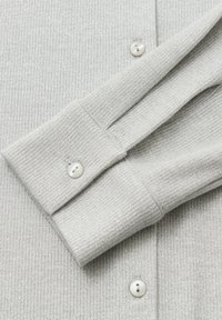 Manica di camicia grigia a coste con una leggera lucentezza, dotata di polsino ripiegato e due bottoni bianchi per l'attacco. Dettaglio del tessuto ad alta texture visibile.