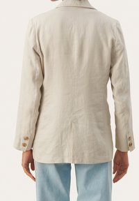 Henkilö, joka on pukeutunut beigeksi pellavablazeriksi, jossa on napitetut hihansuut, ja vaaleansinisiin farkkuihin, kuvattu selkäpuolelta yksinkertaista taustaa vasten.