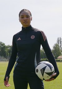 Svartidretts zip-up topp med rosa detaljer og logo, sammen med svarte bukser; holder en svart-hvitt mønstret fotball på gressfelt.