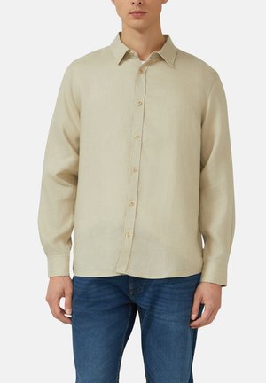 Mann trägt ein beiges, langärmliges Hemd mit Knöpfen und Kragen, kombiniert mit blauen Jeans, steht vor einem schlichten Hintergrund.