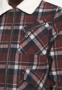 Giacca in flanella a quadri rossi e blu navy con colletto in sherpa bianco, chiusura con cerniera metallica e tasca sul petto con dettaglio a cerniera.