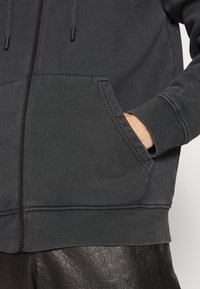 Felpa grigia scura con zip realizzata in tessuto misto cotone. Presenta due tasche frontali e polsini a coste sulle maniche e sull'orlo. Texture liscia.