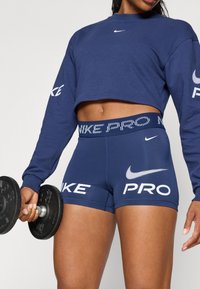 Tengerészkék, rövidített pulcsi hosszú ujjal, és illő tengerészkék rövidnadrág, melyek fehér "Nike" és "PRO" logókkal rendelkeznek, miközben egy súlyzót tart.