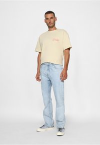 Beige bomullst-shirt med röd text, kombinerad med ljusblå denimjeans och vita sneakers med grå detaljer, stående mot en enfärgad bakgrund.