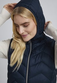 Femme ajustant la capuche d'un gilet matelassé bleu marine porté sur un haut à manches longues crème, regardant vers le bas avec un léger sourire.