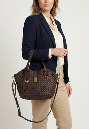 Kvinde iført navy blazer og beige bukser, der holder en mørkebrun fletlæderhåndtaske med guld hardware og skulderrem.