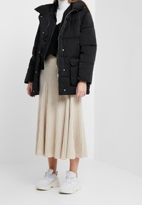 Chaqueta inflable negra con cuello alto, textura acolchada, cierre de cremallera y bolsillos frontales, llevar sobre una blusa negra y una falda de canalé beige.