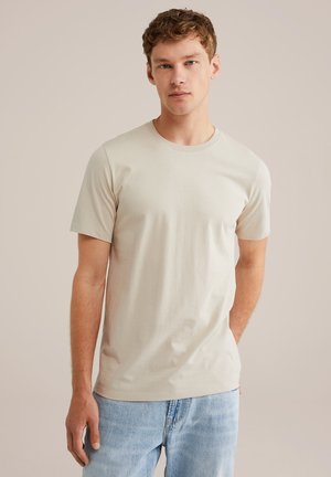T-shirt en coton beige avec un col rond classique et des manches courtes, associé à un jean en denim bleu clair. Texture lisse et design épuré.