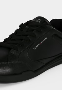 Μαύρο δερμάτινο αθλητικό παπούτσι Tommy Hilfiger με μαύρα κορδόνια και λογότυπο στην πλευρά και τη γλώσσα, σε μονόχρωμο φόντο.