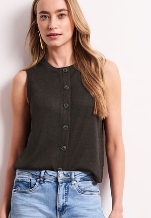 Femme blonde portant un haut sans manches foncé boutonné et un jean bleu clair, debout contre un mur de couleur claire.