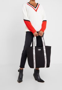 Černá plátěná taška přes rameno s bílým logem a kontrastními proužkovanými rukojetmi. Nositelná s tlustým svetrem s výstřihem do V a červenými akcenty.