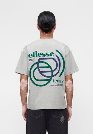 Άτομο με μαλλιά μαζεμένα πίσω, που φοράει γκρι μπλουζάκι με πράσινο και μπλε αφηρημένο σχέδιο τέννις και κείμενο, κοιτάζοντας προς τα πίσω.