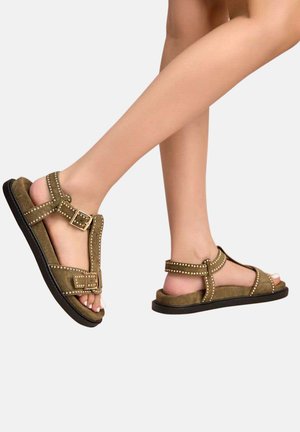 Jambes portant des sandales en daim vert olive avec des clous dorés et des sangles réglables à boucle, présentées sur un fond gris clair.