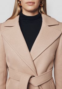 Beżowy wełniany płaszcz z szerokimi klapami i związanym paskiem, noszony na czarnym golfie. Charakteryzuje się prostymi liniami i miękką fakturą.