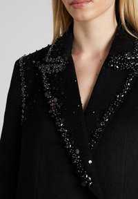 Abrigo de lana negro con un cuello con muescas adornado con lentejuelas y cuentas negras a lo largo de los bordes, creando un detalle texturizado.