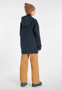 Giacca blu con cappuccio e maniche lunghe, abbinata a pantaloni marroni chiari e stivali neri. Berretto di lana marrone. Tessuto liscio. Nessun motivo visibile.