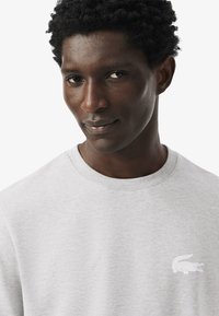 Szürke pamut pulóver, kerek nyakkal, bordás mandzsettával és fehér hímzett krokodil logóval a bal mellrészen. Puha, texturált felület.