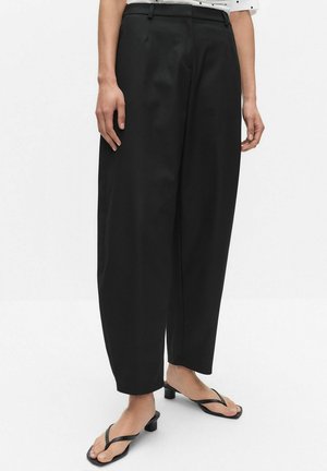 Kvinde iført sorte højtaljede vidde bukser kombineret med sorte hælede tåsandaler, stående på en hvid baggrund.