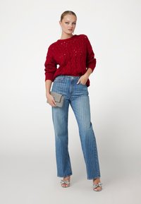 Pull en tricot rouge avec un motif en relief, associé à un jean bleu rayé et des sandales argentées. Le modèle tient une pochette texturée grise.