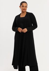 Zwarte lange cardigan met gedrapeerde voorkant, voorzien van lange mouwen met kralenaccenten. Gemaakt van zachte stof, ontworpen voor comfort.