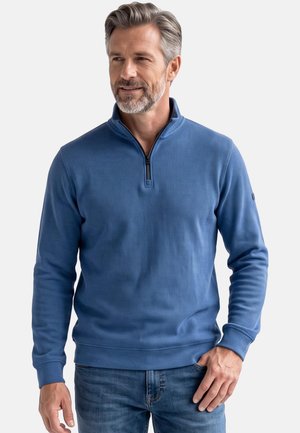 Mann mit grauen Haaren und Bart, der einen blauen Pullover mit Viertelreißverschluss und Jeans trägt, vor einem schlichten hellen Hintergrund steht.