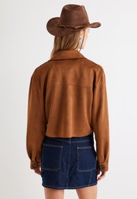 Veste cropped en daim marron avec un col, associée à une jupe en denim bleu foncé avec deux poches arrière. Chapeau marron avec un bandana décoratif.