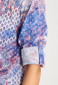 Long-sleeve shirt featuring a blue and pink paisley pattern, rolled cuffs, and contrasting button detail. Made from a lightweight, textured fabric.
