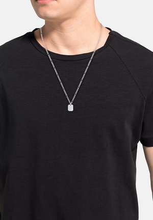Collana d'argento con un pendente rettangolare; presenta una superficie testurizzata. Indossata sopra una maglietta nera a maniche corte.
