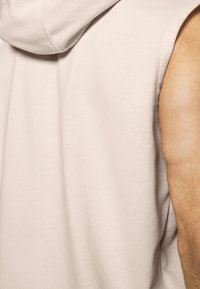 Light beige sleeveless hoodie with a smooth texture, featuring a hood and rounded edges, highlighting muscular arm detail.