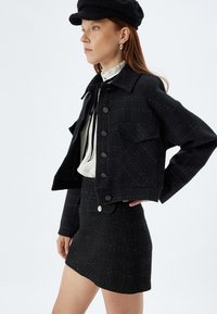Ensemble composé d'une veste et d'une jupe en tweed noir avec un motif texturé, présentant de grands boutons, des poches latérales et un design de veste courte.