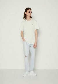 Ανοιχτό πράσινο βαμβακερό t-shirt με μικρό λογότυπο, σε συνδυασμό με ανοιχτό μπλε σκισμένα τζιν και λευκά αθλητικά παπούτσια. Το μοντέλο φορά καφέ γυαλιά ηλίου.